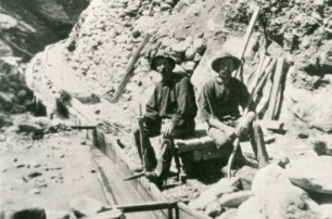 Two gold miners wearing hats resting alongside pickaxes and mining pans